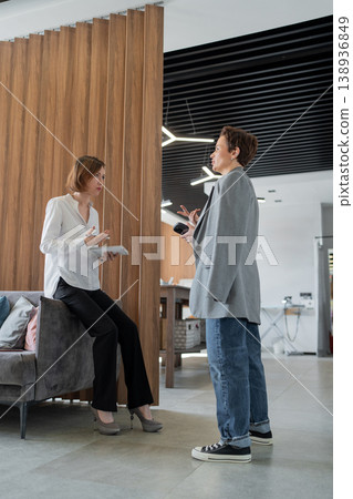 Two women chatting and gesturing while standing near a wooden wall in a modern office. Chatting, gestures, teamwork, standing, office. 138936849
