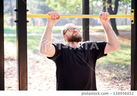 White bearded man concentrates during workout. Novice trainer with beard performing exercise under sunny trees. Bearded man in black shirt is focused on lifting weights outdoors amidst sunlit trees 138936852