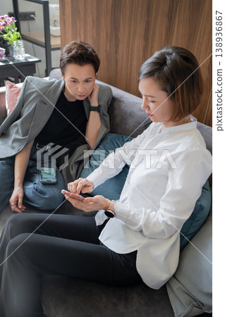Confident women in business clothes talking and using smartphone together on couch in office. Confidence, smartphone, business, teamwork, office. 138936867