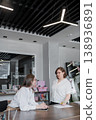 Women standing across table from each other in office, business discussion, paperwork and folders present. Business, paperwork, table, discussion, women, folders. 138936891