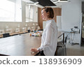 Businesswoman standing and speaking in bright open office area with desk near large windows. Speaking, office, desk, windows, businesswoman, modern. 138936909