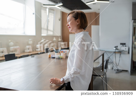 Businesswoman standing and speaking in bright open office area with desk near large windows. Speaking, office, desk, windows, businesswoman, modern. 138936909