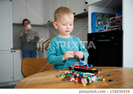 Boy playing with construction blocks. Sitting at table at home, building toy model with colorful pieces, mother in background drinking coffee in kitchen 138936956