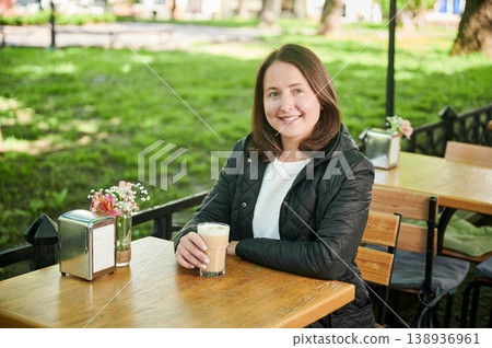 Smiling woman sitting at outdoor cafe table holding glass of latte, enjoying peaceful moment in park setting, natural light, casual lifestyle portrait Smiling woman sitting at outdoor cafe table holding glass of latte, enjoying peaceful moment in park setting, natural light, casual lifestyle portrait 138936961