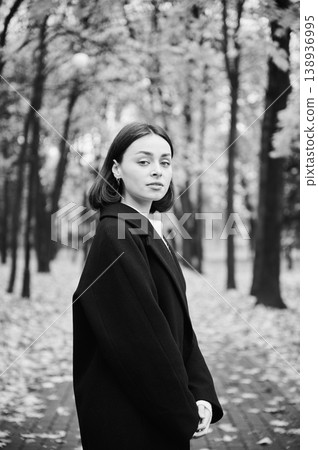 Young woman posing in black coat on park path in monochrome autumn portrait, looking at camera among blurred trees. Ideal for fashion, beauty, timeless style, and advertising Young woman posing in black coat on park path in monochrome autumn portrait, looking at camera among blurred trees. Ideal for fashion, beauty, timeless style, and advertising 138936995