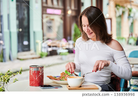 Smiling woman eating meal and drinking coffee at outdoor cafe, enjoying relaxed breakfast or brunch, urban lifestyle and leisure time concept Smiling woman eating meal and drinking coffee at outdoor cafe, enjoying relaxed breakfast or brunch, urban lifestyle and leisure time concept 138936998