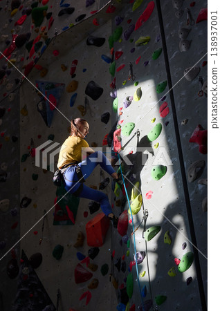 Female climber ascends artificial rock wall with strong shadow and spotlight in sports gym. Great for determination, endurance, motivation, recreation or indoor sport concepts Female climber ascends artificial rock wall with strong shadow and spotlight in sports gym. Great for determination, endurance, motivation, recreation or indoor sport concepts 138937001
