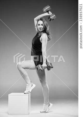 Black and white studio shot of athletic woman holding aquafitness dumbbells with foot raised on cube, smiling at camera. Dynamic pose. Perfect for water fitness, equipment, and active lifestyle ads Black and white studio shot of athletic woman holding aquafitness dumbbells with foot raised on cube, smiling at camera. Dynamic pose. Perfect for water fitness, equipment, and active lifestyle ads 138937021