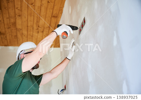 Electrician installing wall outlet with drill and securing electrical system components. Worker in helmet performing interior finishing, symbol of home improvement and modern housing development 138937025