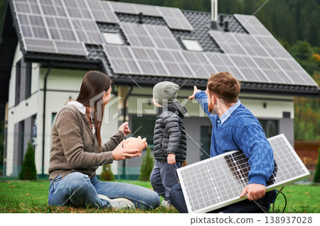 Father pointing at solar panels while mother holds piggy bank and child stands near modern house. Photovoltaic rooftop system, renewable energy, saving concept, eco family lifestyle Father pointing at solar panels while mother holds piggy bank and child stands near modern house. Photovoltaic rooftop system, renewable energy, saving concept, eco family lifestyle 138937028