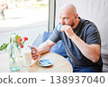 Man drinking coffee and using smartphone in cafe by window, holding cup and checking phone at table with flowers and water, modern lifestyle and digital communication concept 138937040
