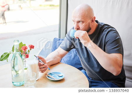 Man drinking coffee and using smartphone in cafe by window, holding cup and checking phone at table with flowers and water, modern lifestyle and digital communication concept Man drinking coffee and using smartphone in cafe by window, holding cup and checking phone at table with flowers and water, modern lifestyle and digital communication concept 138937040