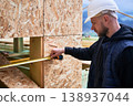 Carpenter measuring horizontal distance across wooden facade cutout, verifying alignment and installation accuracy on building site 138937044