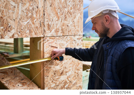 Carpenter measuring horizontal distance across wooden facade cutout, verifying alignment and installation accuracy on building site Carpenter measuring horizontal distance across wooden facade cutout, verifying alignment and installation accuracy on building site 138937044