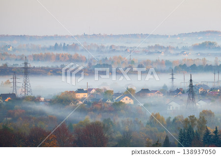 Fog drifts over suburban houses and autumn forest in layered morning landscape with power lines. Suitable for weather, environment, energy infrastructure, rural living or seasonal themes Fog drifts over suburban houses and autumn forest in layered morning landscape with power lines. Suitable for weather, environment, energy infrastructure, rural living or seasonal themes 138937050
