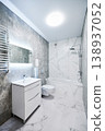 Contemporary bathroom interior with bathtub, suspended toilet and sink cabinet. Refined washroom with cool gray stone texture, white marble wall panels, backlit mirror and towel radiator 138937052