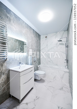 Contemporary bathroom interior with bathtub, suspended toilet and sink cabinet. Refined washroom with cool gray stone texture, white marble wall panels, backlit mirror and towel radiator 138937052