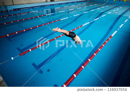 Male swimmer entering water after dive during sprint start. Athlete in streamlined position breaking surface, indoor swimming pool lanes, fast competition and training action concept 138937058