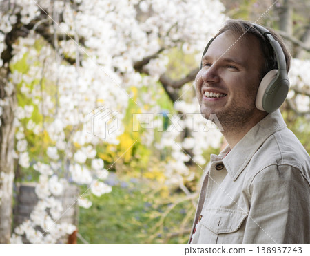 A smiling man wearing headphones enjoys music among blooming spring trees in a Japanese park, creating a cinematic backdrop. 138937243