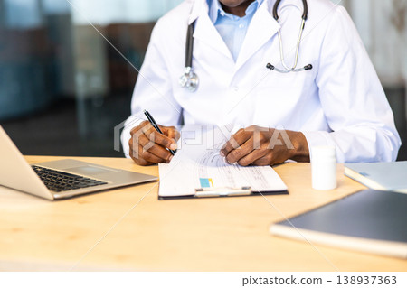 Medical professional in white coat with stethoscope reviews patient files and health records while working on a laptop at a wooden desk, managing documentation and care planning Medical professional in white coat with stethoscope reviews patient files and health records while working on a laptop at a wooden desk, managing documentation and care planning 138937363