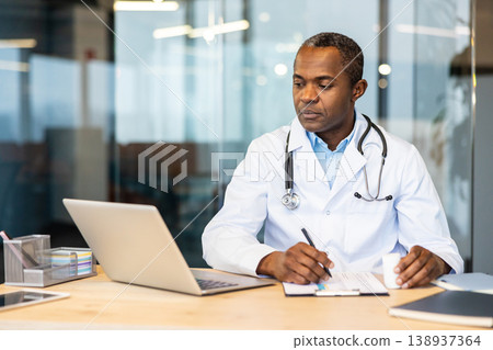 Experienced physician focused at a wooden desk with laptop and documents, reviewing patient records and providing professional medical consultation and diagnostic support in a clinic office Experienced physician focused at a wooden desk with laptop and documents, reviewing patient records and providing professional medical consultation and diagnostic support in a clinic office 138937364