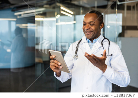 Experienced physician wearing a white lab coat and stethoscope, smiling while holding a digital tablet and gesturing, conducting an online medical consultation from a modern office environment Experienced physician wearing a white lab coat and stethoscope, smiling while holding a digital tablet and gesturing, conducting an online medical consultation from a modern office environment 138937367