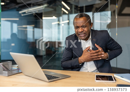 Mature businessman experiencing sudden, severe chest pain and heart discomfort while working in a modern office, symbolizing workplace stress, health crises, and the need for medical emergency Mature businessman experiencing sudden, severe chest pain and heart discomfort while working in a modern office, symbolizing workplace stress, health crises, and the need for medical emergency 138937373