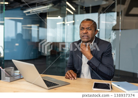 Mature businessman experiencing pain and discomfort in his throat while working on a laptop at his office desk, indicating illness and struggling with health issues during work hours Mature businessman experiencing pain and discomfort in his throat while working on a laptop at his office desk, indicating illness and struggling with health issues during work hours 138937374