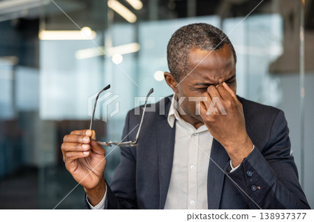 Businessman enduring stress from work, experiencing eye strain and a headache, rubbing his eyes while holding his spectacles indoors at the modern office Businessman enduring stress from work, experiencing eye strain and a headache, rubbing his eyes while holding his spectacles indoors at the modern office 138937377