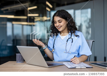 Young female healthcare provider wearing scrubs and stethoscope sitting at desk, having a virtual meeting or teleconsultation with a patient, writing notes while communicating online 138937393