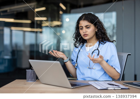 Young indian female doctor wearing scrubs and a stethoscope, showing confusion and concern while having a video call on a laptop in a modern office, expressing frustration Young indian female doctor wearing scrubs and a stethoscope, showing confusion and concern while having a video call on a laptop in a modern office, expressing frustration 138937394