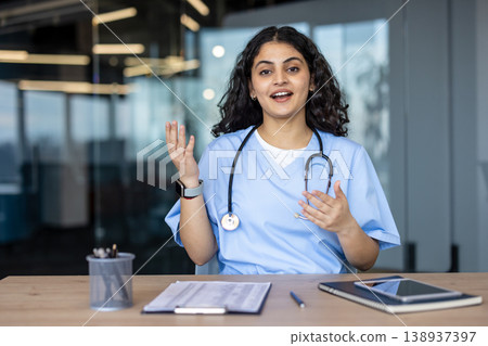 Young indian female doctor wearing scrubs and a stethoscope, gesturing while engaging in a professional online remote medical consultation from a modern office environment Young indian female doctor wearing scrubs and a stethoscope, gesturing while engaging in a professional online remote medical consultation from a modern office environment 138937397