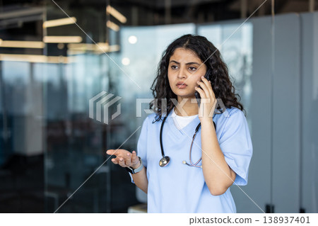 Female medical professional in scrubs and a stethoscope talking on a mobile phone, actively communicating while gesturing, standing in a modern clinic or hospital setting Female medical professional in scrubs and a stethoscope talking on a mobile phone, actively communicating while gesturing, standing in a modern clinic or hospital setting 138937401