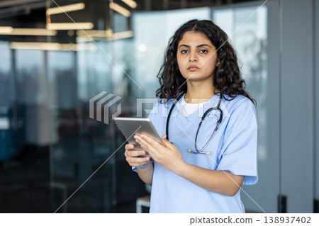 Young female medical professional in scrubs and stethoscope standing while holding a digital tablet, representing modern healthcare, medical technology, and dedicated patient care 138937402