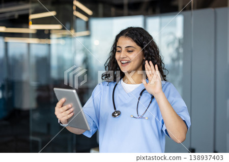 Young woman nurse or doctor wearing scrubs and stethoscope, engaging in a virtual consultation with a patient via video call on a digital tablet, providing telehealth services 138937403