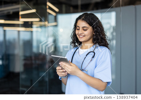 Young female nurse in light-blue scrubs and stethoscope smiling while using a tablet for patient records and telehealth in a modern clinic, showcasing digital healthcare integration 138937404