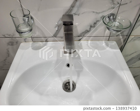 Water flows from the tap in the bathroom, where there is a white handwashing sink with glass accessories Water flows from the tap in the bathroom, where there is a white handwashing sink with glass accessories 138937410