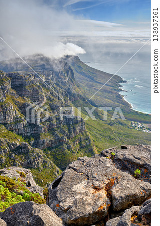 Clouds and fog covering a mountain peak on a sunny day along the coast. Scenic landscape with beautiful views of a rocky terrain in nature. Calm sea around an iconic landmark and travel destination Clouds and fog covering a mountain peak on a sunny day along the coast. Scenic landscape with beautiful views of a rocky terrain in nature. Calm sea around an iconic landmark and travel destination 138937561