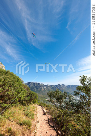 Nature park landscape with plants and an outdoor dirt path. Beautiful outdoors view of a hiking trail in the mountains during summer. Mountain background with a blue sky and paragliding travelers. 138937588