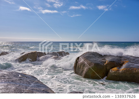 Beautiful landscape view of the rough ocean waves breaking on large boulders with a blue sky background. Tidal waves crashing into rocks on the windy wild coast and beach of Cape Town, South Africa Beautiful landscape view of the rough ocean waves breaking on large boulders with a blue sky background. Tidal waves crashing into rocks on the windy wild coast and beach of Cape Town, South Africa 138937626