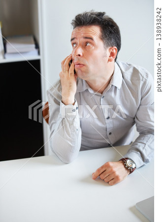 Phone call, thinking or waiting with a business man sitting at his desk in the office for work. Time, contact and communication with an impatient young employee talking on his mobile in the workplace Phone call, thinking or waiting with a business man sitting at his desk in the office for work. Time, contact and communication with an impatient young employee talking on his mobile in the workplace 138938242