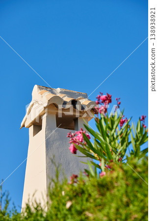Chimney tower designed roof house building outside against blue sky background and green plant foreground. Construction of exterior architecture of chute built on rooftop for fireplace smoke and heat Chimney tower designed roof house building outside against blue sky background and green plant foreground. Construction of exterior architecture of chute built on rooftop for fireplace smoke and heat 138938921