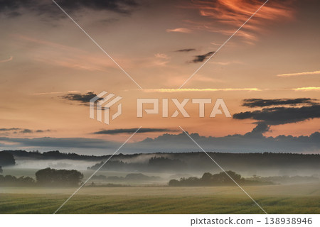 Beautiful sunrise with mist against a dramatic and colorful sky background. Calm and breathtaking landscape view on a field in the countryside with blooming trees on a foggy and overcast morning 138938946