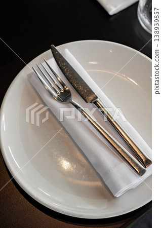Silver fork and knife placed on white napkin inside round plate on dark table setting Silver fork and knife placed on white napkin inside round plate on dark table setting 138939857