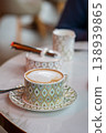 Decorative coffee cup with frothy cappuccino on patterned saucer, sugar bowl and milk jug nearby 138939865