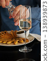Person seasoning fried food with salt while holding a glass of white wine at a dining table 138939873