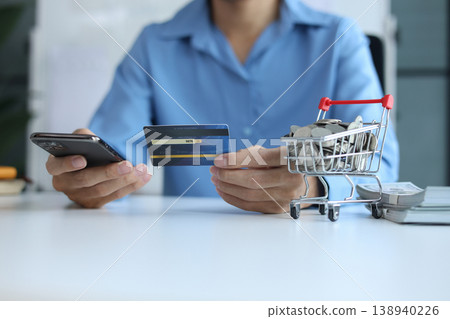 Person using smartphone and credit card for online shopping with stack of cash and coins in shopping cart. 138940226