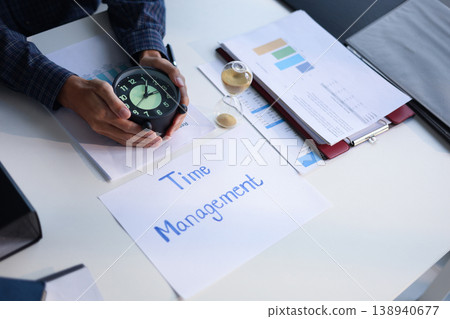 Time management concept. Businessman holding clock while working on office desk with stack of documents on the table. Time management concept. Businessman holding clock while working on office desk with stack of documents on the table. 138940677