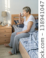 Senior woman taking a drink of water in her bedroom, sitting on the bed. Practicing her daily health and wellness routine, medication routine, prevention of age-related diseases 138941047