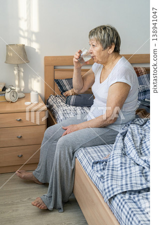 Senior woman taking a drink of water in her bedroom, sitting on the bed. Practicing her daily health and wellness routine, medication routine, prevention of age-related diseases 138941047